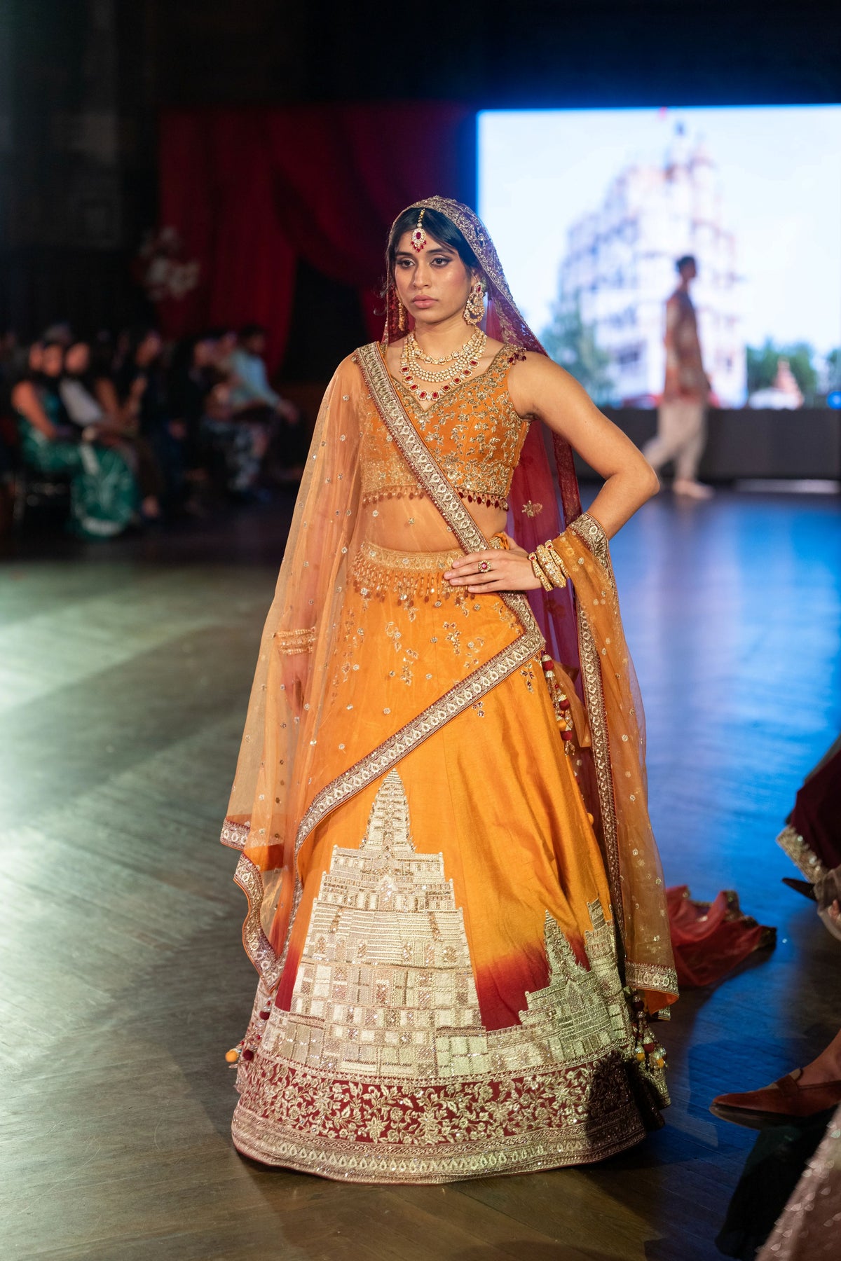 Angkor Wat orange lehenga with a temple-architecture panel at the hem, model standing with a hand on her hip