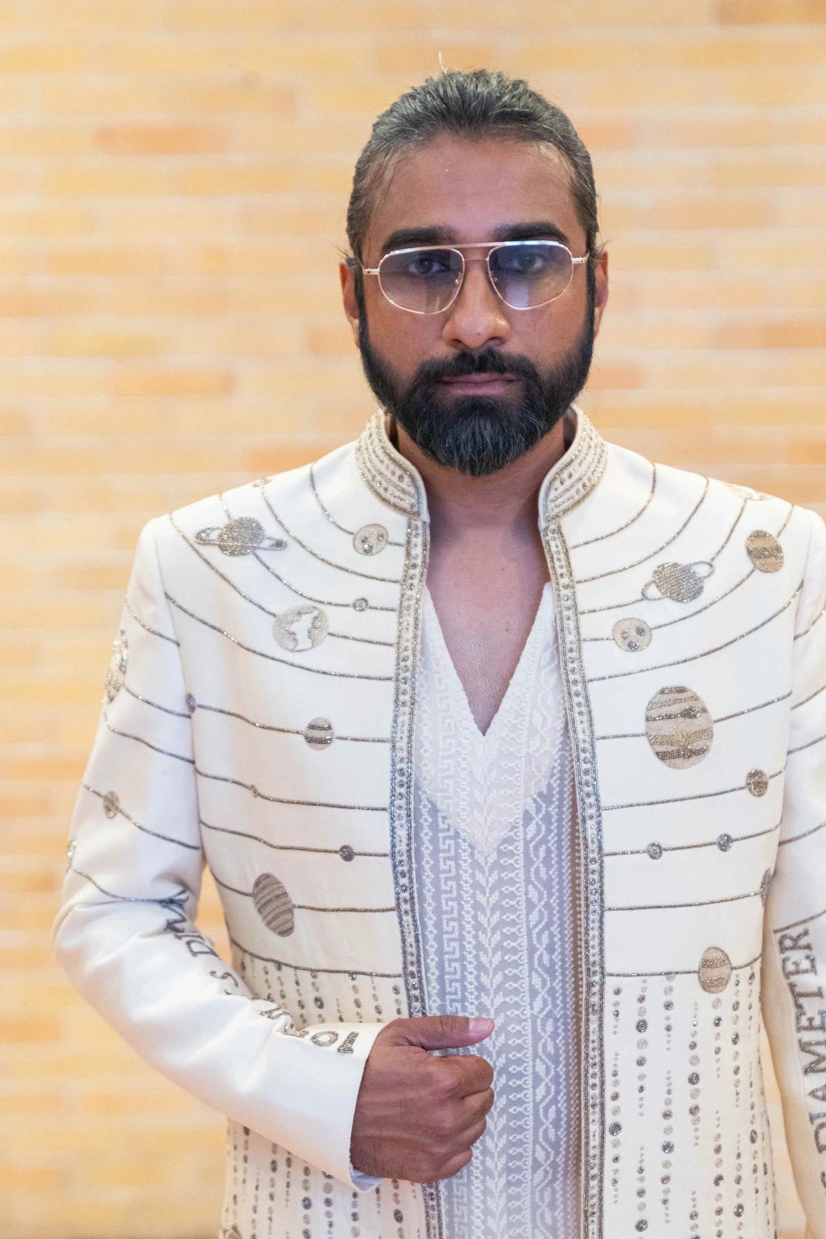 Close-up front of men's ivory Hanuman Chalisa jacket with planetary orbit zardozi lines and discs against a brick wall