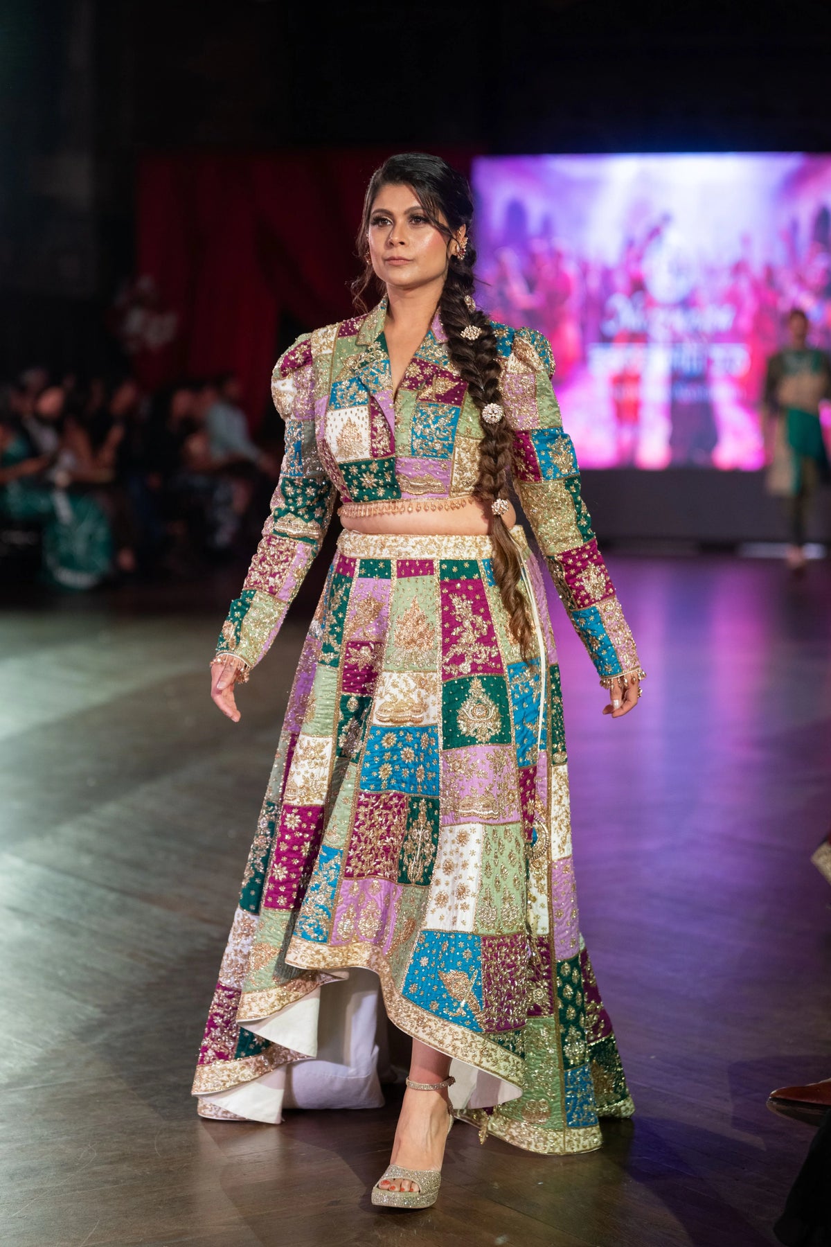 Closer runway view of the Holi patchwork lehenga with a long braid and high-low hem under a screen backdrop