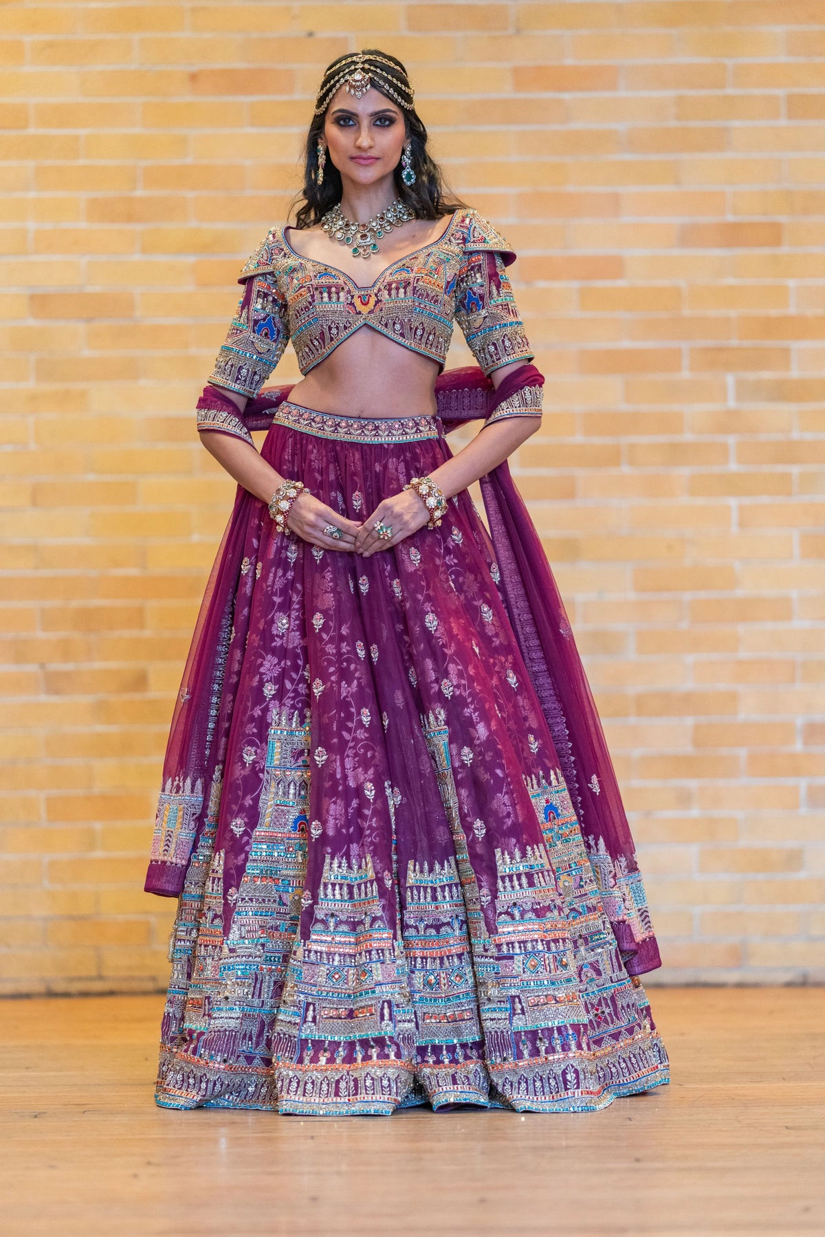 Front view of the Meenakshi Temple magenta lehenga with a multicolor gopuram-pattern hem, model standing against a brick wall