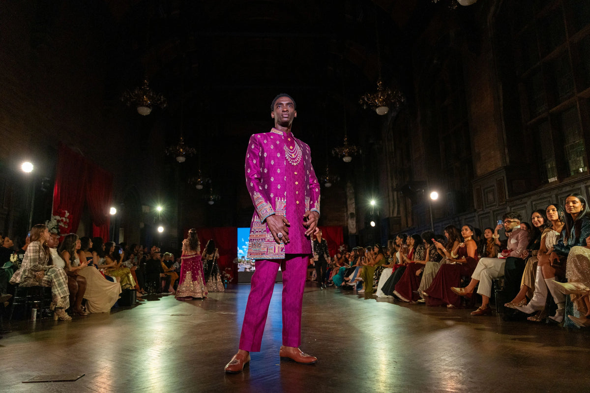 Man in a magenta Meenakshi Temple open sherwani with gold embroidery standing on the runway, audience seated along the side