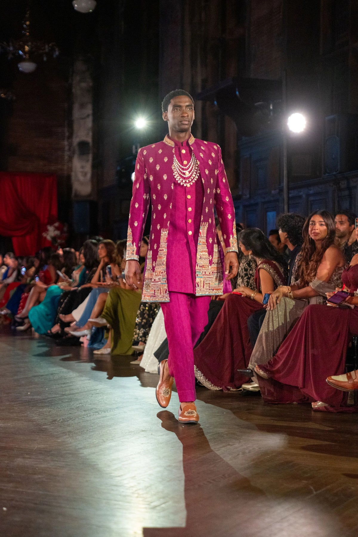 Close three-quarter view of the magenta sherwani with gold booti and embroidered hem, audience seated behind