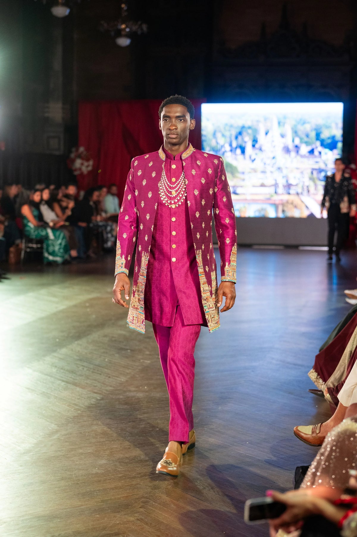 Model walking toward the camera in the magenta Meenakshi Temple sherwani and kurta with a runway screen behind
