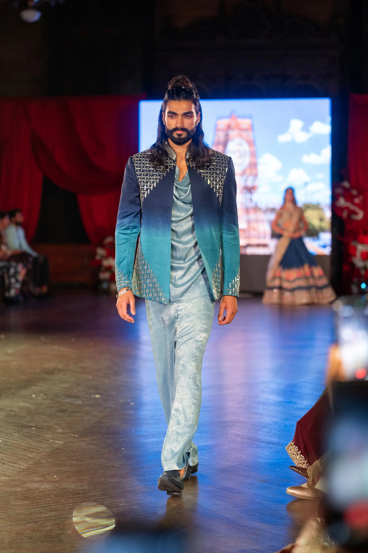 Model walking the runway in the Meru Prastara blue ombre jacket with pale blue kurta and trousers, temple image on screen
