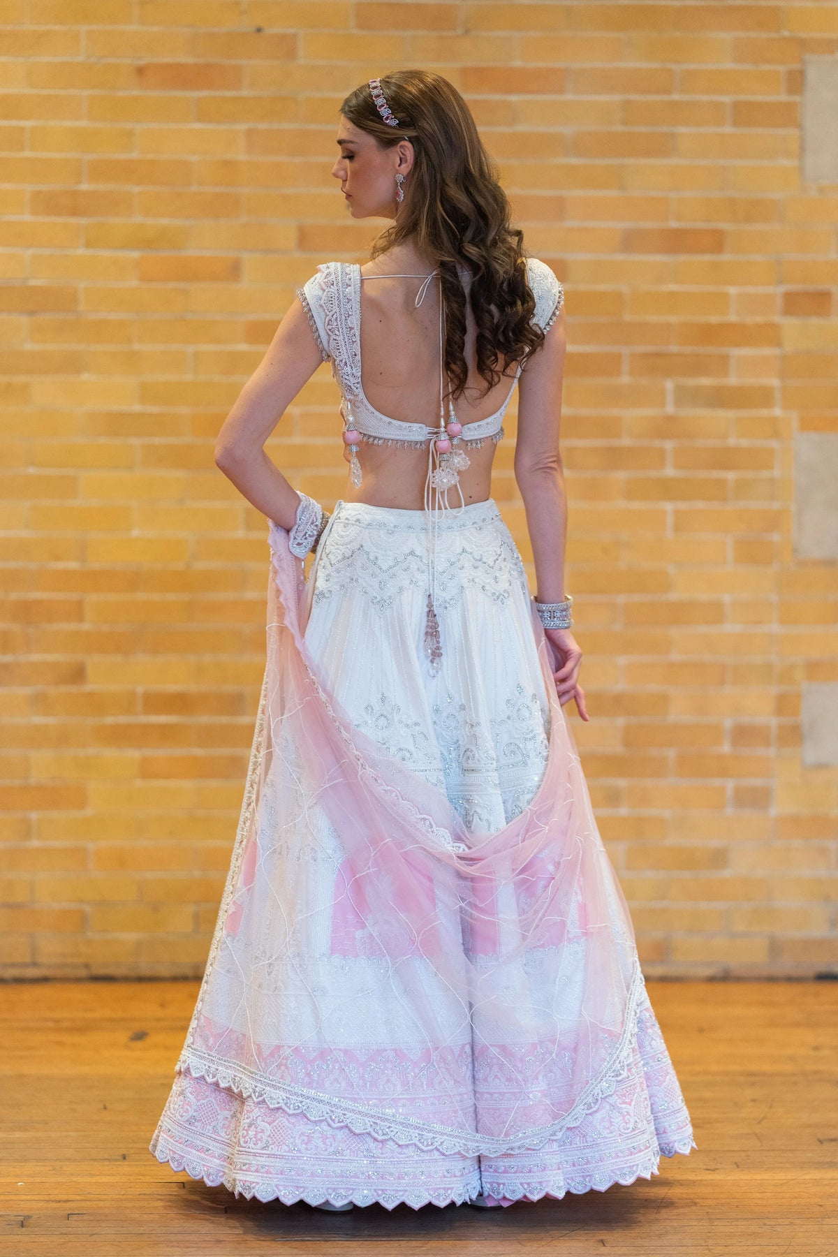 Back view of the Prem Mandir lehenga with an open-back embroidered blouse and a pink dupatta against a brick wall