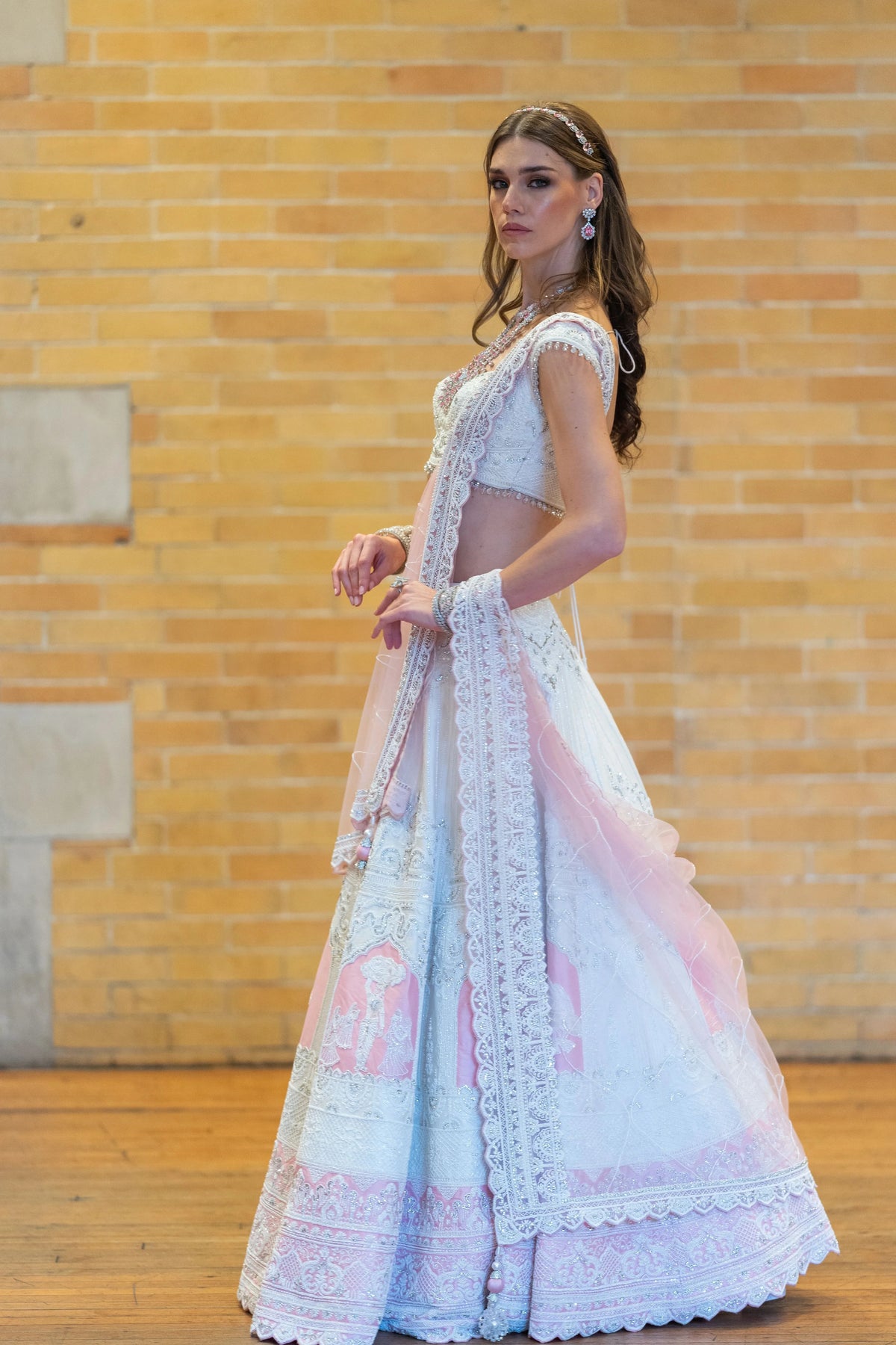 Side pose in the Prem Mandir pale lehenga with pink floral panels and lace hem against a brick wall