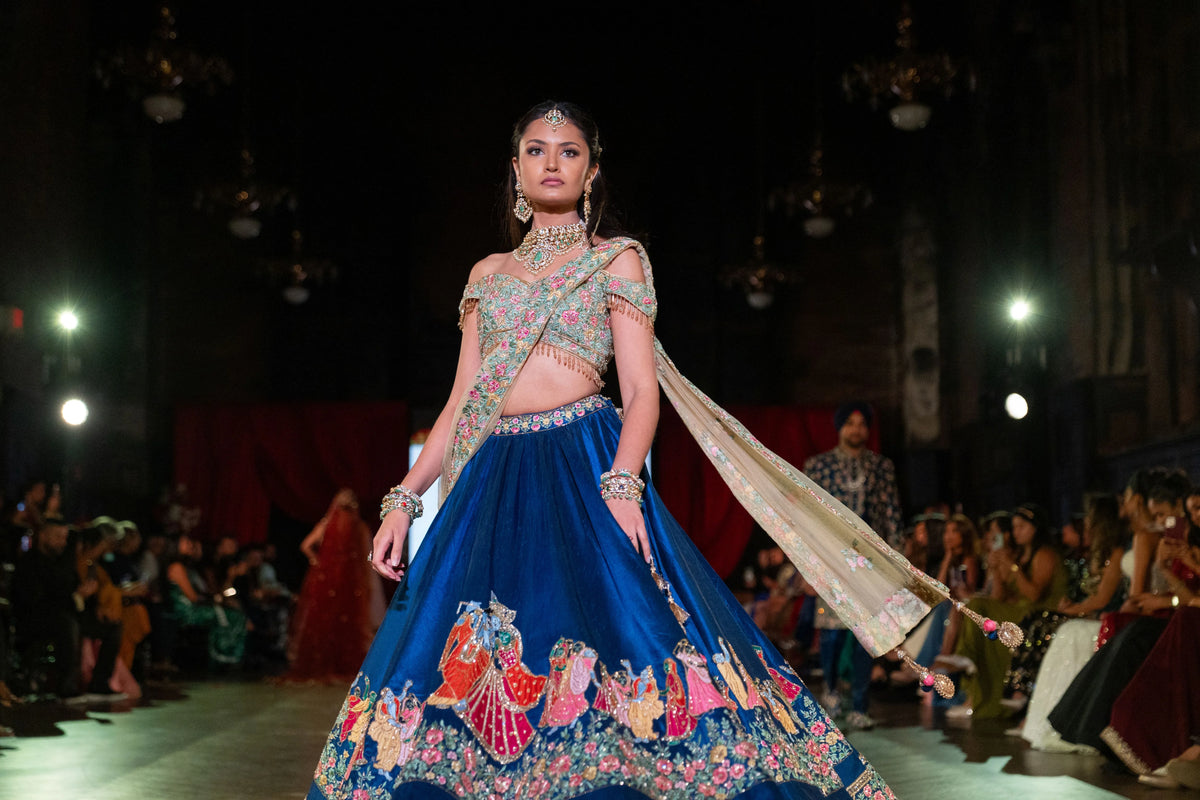 Wide runway view of the Raas Leela blue lehenga set with seated guests and a temple image on screen