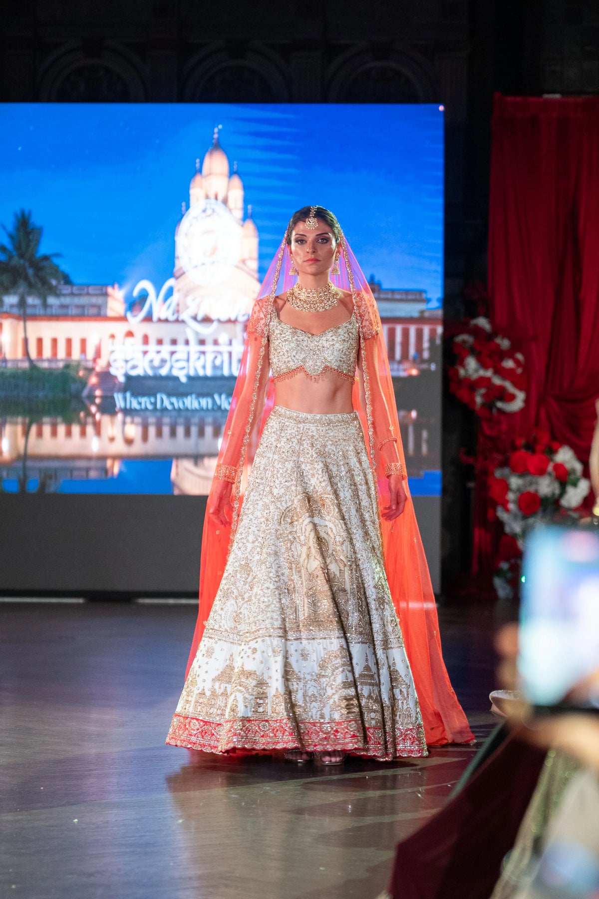 Model in the Siddhivinayak ivory lehenga with a red embroidered border and orange-red dupatta, temple image on the screen behind
